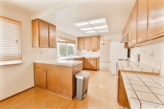 kitchen with tile countertops, light wood-style flooring, a peninsula, a chandelier, and under cabinet range hood