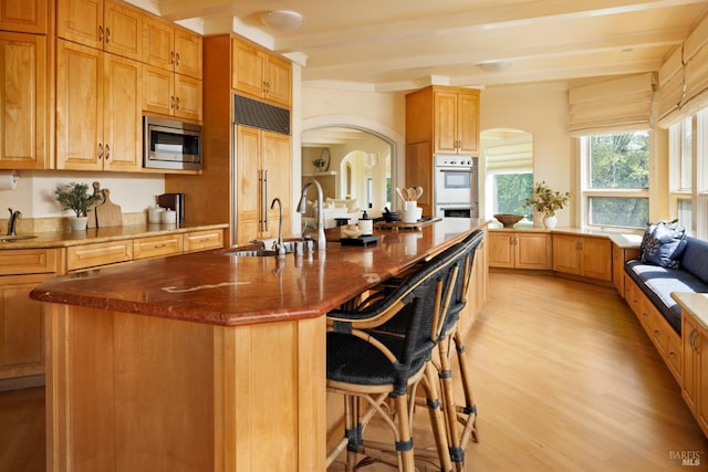 kitchen featuring built in appliances, light wood-style floors, a kitchen breakfast bar, a center island with sink, and arched walkways