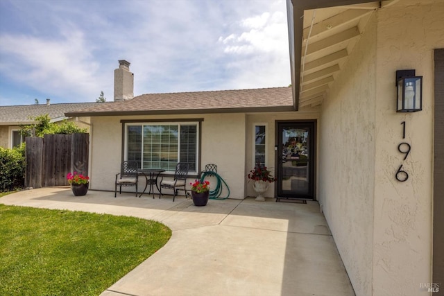 view of exterior entry with a patio area, fence, a yard, stucco siding, and a shingled roof