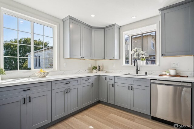kitchen with stainless steel dishwasher, gray cabinetry, a sink, light wood-style floors, and light stone countertops