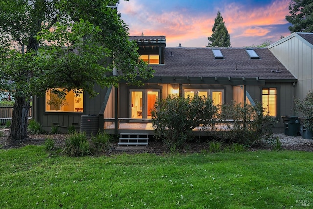 rear view of house with a yard, a deck, a shingled roof, and central AC