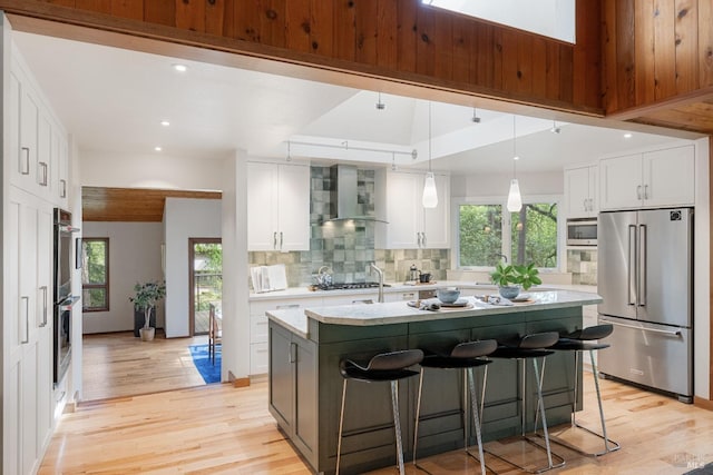 kitchen with wall chimney range hood, decorative backsplash, light wood-type flooring, and appliances with stainless steel finishes