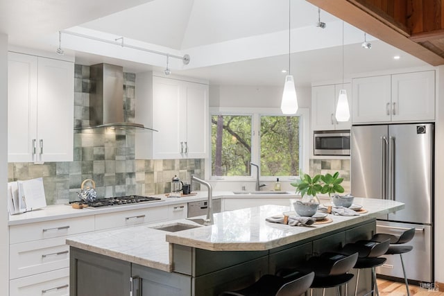 kitchen with a sink, wall chimney exhaust hood, backsplash, stainless steel appliances, and an island with sink