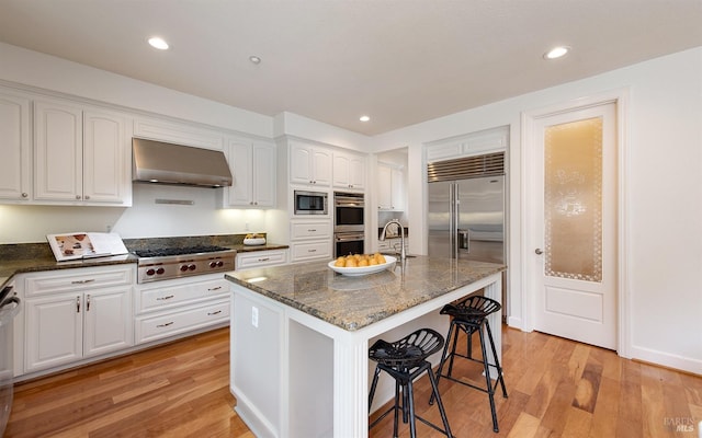 kitchen with dark stone counters, wall chimney exhaust hood, built in appliances, and light wood-style floors