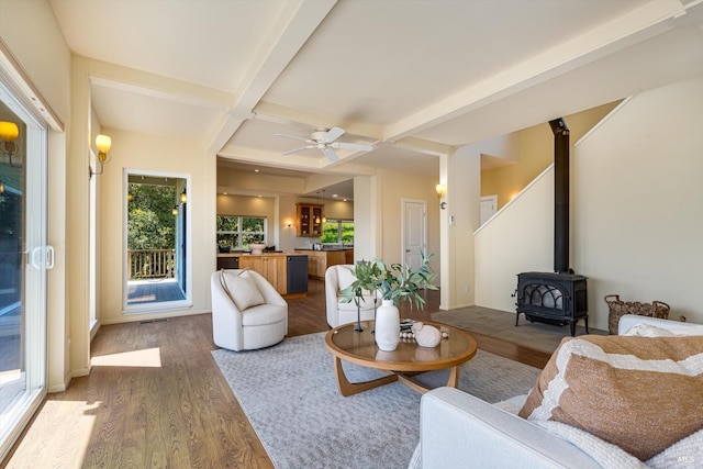 living room with a wood stove, light wood-style flooring, ceiling fan, and beamed ceiling