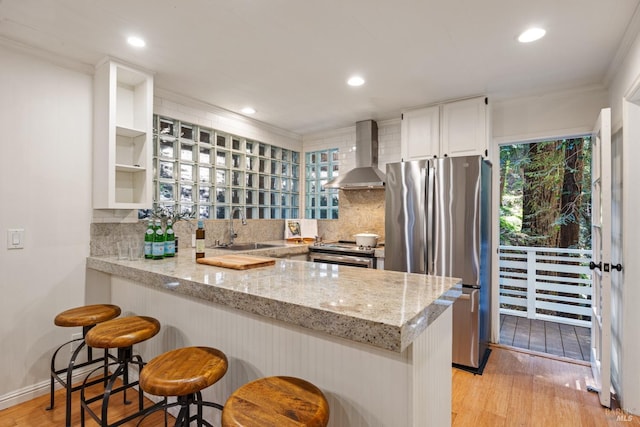 kitchen featuring a sink, a peninsula, wall chimney exhaust hood, backsplash, and stainless steel appliances