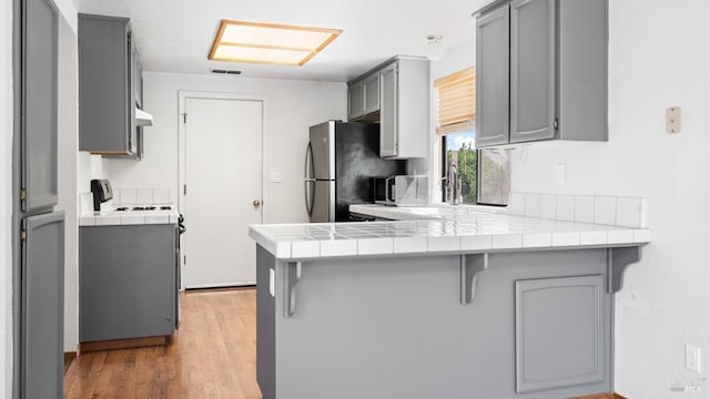 kitchen featuring gray cabinets, a peninsula, appliances with stainless steel finishes, tile counters, and light wood-type flooring