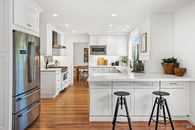kitchen featuring appliances with stainless steel finishes, wall chimney range hood, a peninsula, decorative backsplash, and white cabinetry