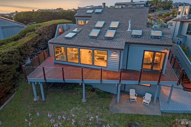 back of property at dusk with roof with shingles, a deck, and a patio area