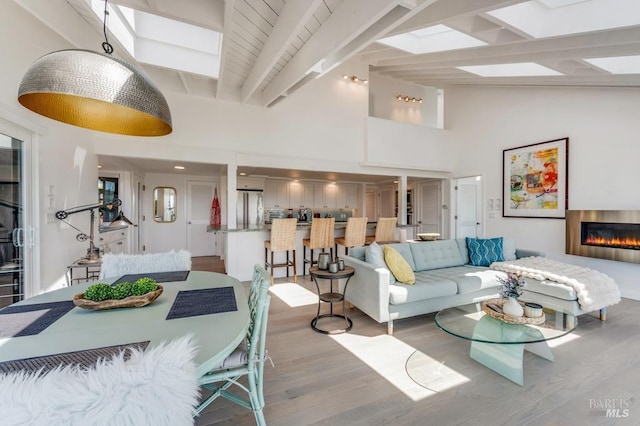 living area featuring wood finished floors, a skylight, beamed ceiling, a glass covered fireplace, and high vaulted ceiling