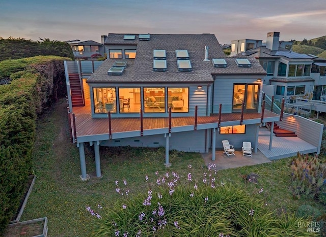 back of house at dusk featuring a wooden deck, roof with shingles, and a patio area