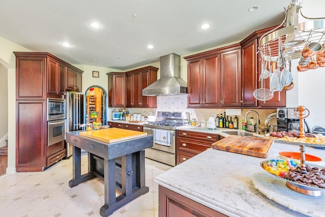 kitchen with wall chimney exhaust hood, appliances with stainless steel finishes, a sink, reddish brown cabinets, and tasteful backsplash