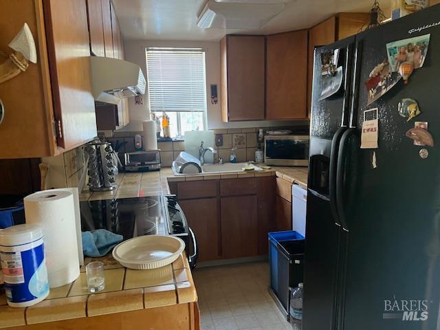 13238 Ebbtide Vlg, Clearlake Oaks, CA 95423 - kitchen with black fridge with ice dispenser, tile counters, exhaust hood, and brown cabinetry