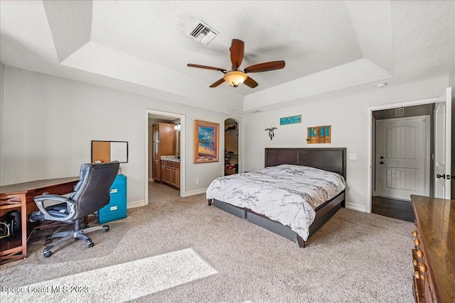 bedroom with a tray ceiling, ceiling fan, ensuite bathroom, and light colored carpet