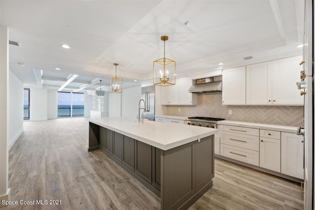 kitchen with an island with sink, light hardwood / wood-style floors, wall chimney range hood, tasteful backsplash, and white cabinetry