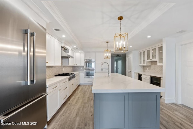 kitchen featuring appliances with stainless steel finishes, white cabinets, a center island with sink, wall chimney range hood, and tasteful backsplash