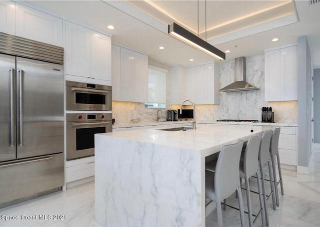 kitchen featuring backsplash, a center island with sink, stainless steel appliances, wall chimney exhaust hood, and white cabinetry