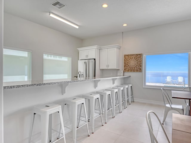 kitchen featuring stainless steel fridge, light tile flooring, a kitchen breakfast bar, white cabinets, and light stone counters