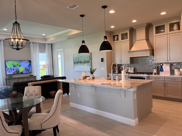 kitchen featuring decorative backsplash, a center island with sink, custom range hood, a notable chandelier, and light stone counters