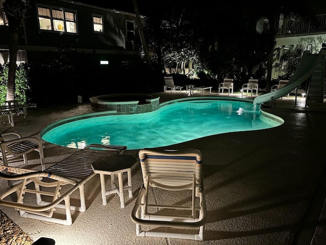 pool at night featuring a water slide, an in ground hot tub, and a patio