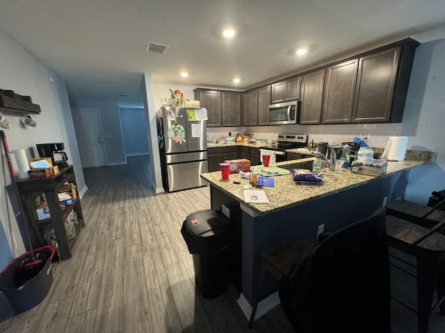 kitchen with stainless steel appliances, light hardwood / wood-style flooring, backsplash, dark brown cabinetry, and light stone counters