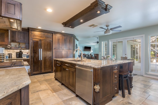 kitchen featuring a kitchen island with sink, paneled built in fridge, dark brown cabinets, stainless steel dishwasher, and ceiling fan