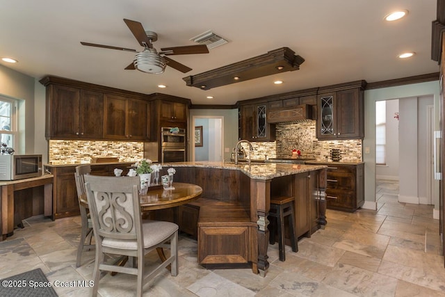 kitchen featuring light stone counters, appliances with stainless steel finishes, range hood, an island with sink, and glass insert cabinets