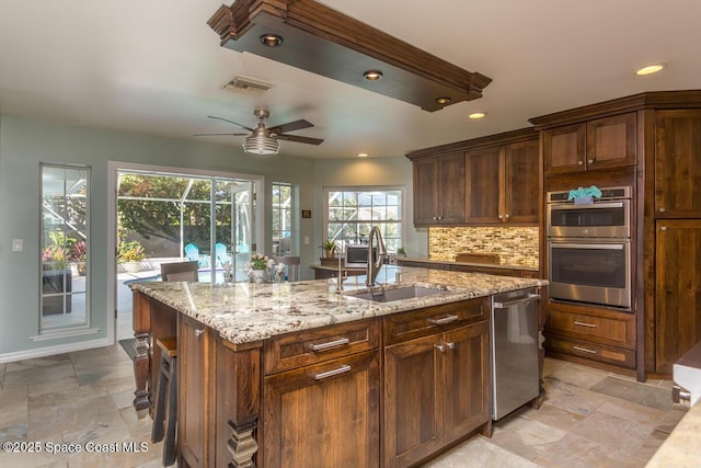 kitchen featuring tasteful backsplash, appliances with stainless steel finishes, light stone countertops, a kitchen island with sink, and a sink