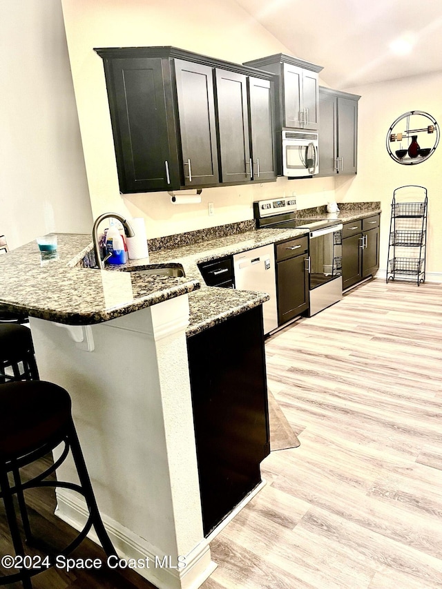 kitchen with kitchen peninsula, appliances with stainless steel finishes, light wood-type flooring, a kitchen breakfast bar, and dark stone countertops