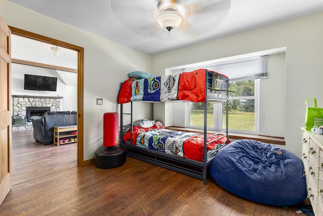 bedroom featuring a stone fireplace, dark wood-type flooring, and ceiling fan