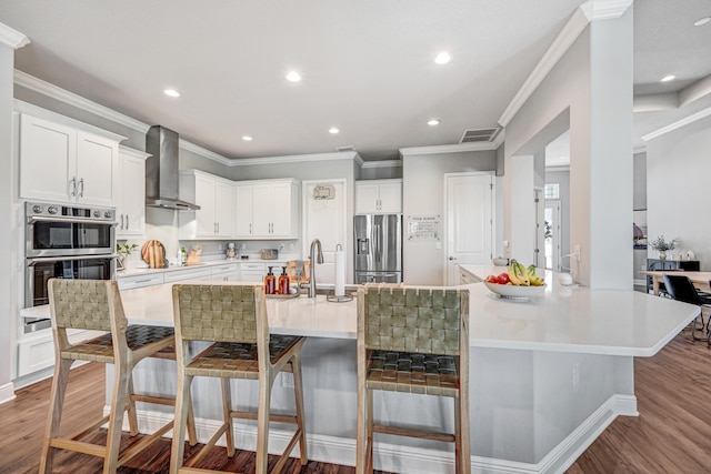 kitchen featuring wood-type flooring, wall chimney range hood, ornamental molding, a kitchen bar, and appliances with stainless steel finishes