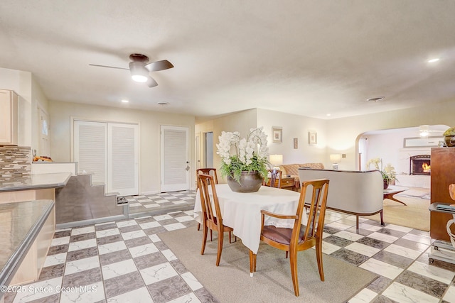 dining space featuring arched walkways, a warm lit fireplace, ceiling fan, and light floors