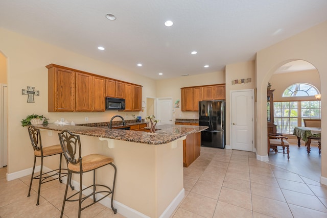 kitchen featuring stainless steel fridge, dark stone counters, sink, light tile patterned floors, and a breakfast bar area