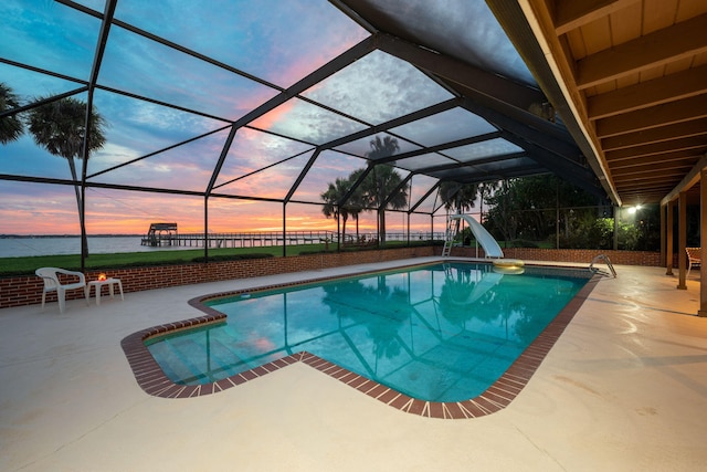 pool at dusk with a water view, a water slide, glass enclosure, and a patio area