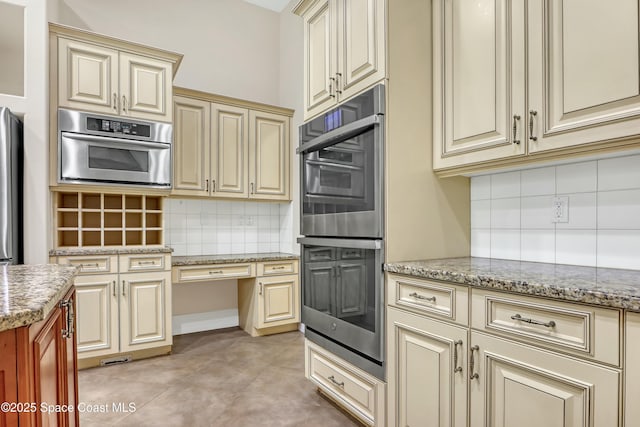 kitchen with appliances with stainless steel finishes, backsplash, light tile patterned floors, light stone countertops, and cream cabinetry