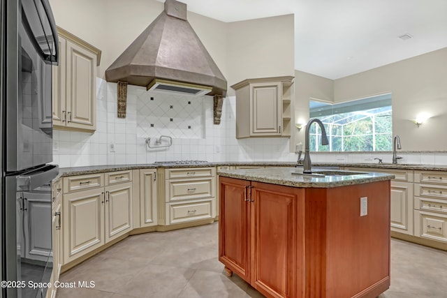 kitchen featuring sink, custom exhaust hood, a center island with sink, stainless steel gas stovetop, and light stone countertops