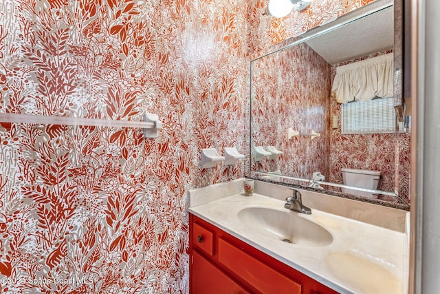 half bath with vanity, a textured ceiling, toilet, and wallpapered walls