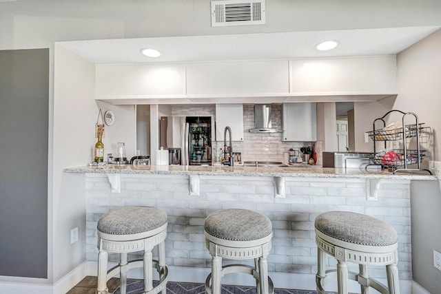 kitchen with kitchen peninsula, light stone counters, a kitchen breakfast bar, and wall chimney exhaust hood