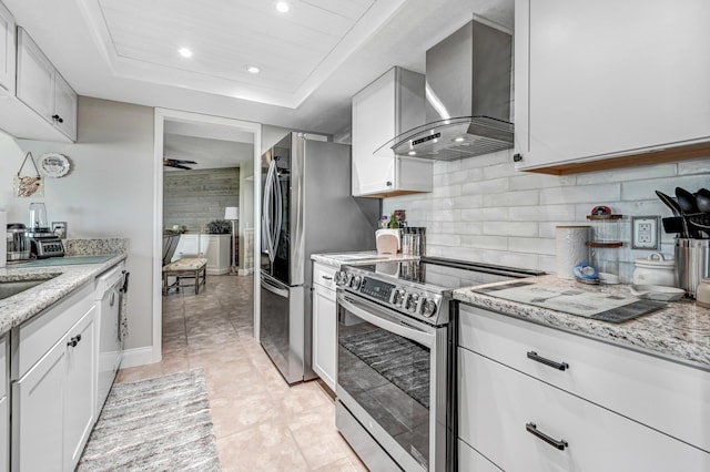 kitchen featuring wall chimney range hood, backsplash, stainless steel appliances, white cabinets, and a raised ceiling