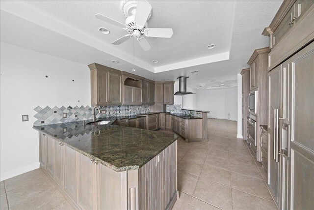 kitchen featuring dark stone countertops, a raised ceiling, kitchen peninsula, island exhaust hood, and ceiling fan