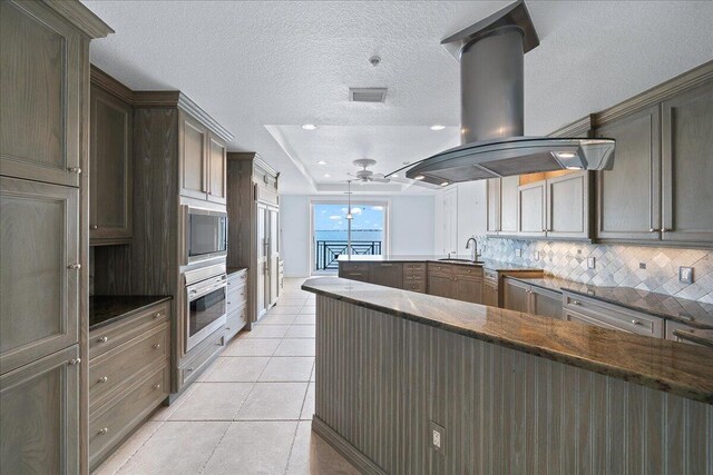 kitchen with dark stone countertops, appliances with stainless steel finishes, island exhaust hood, ceiling fan, and light tile patterned flooring