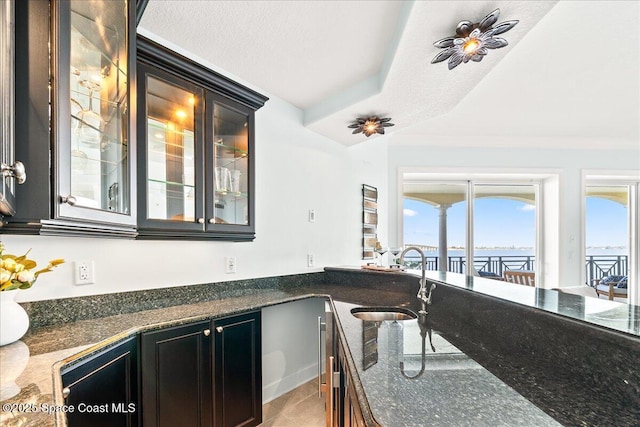 kitchen with a textured ceiling, tile patterned flooring, a sink, dark stone counters, and glass insert cabinets