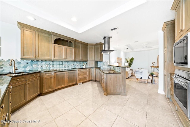 kitchen featuring island exhaust hood, a raised ceiling, dark stone countertops, built in microwave, and a peninsula