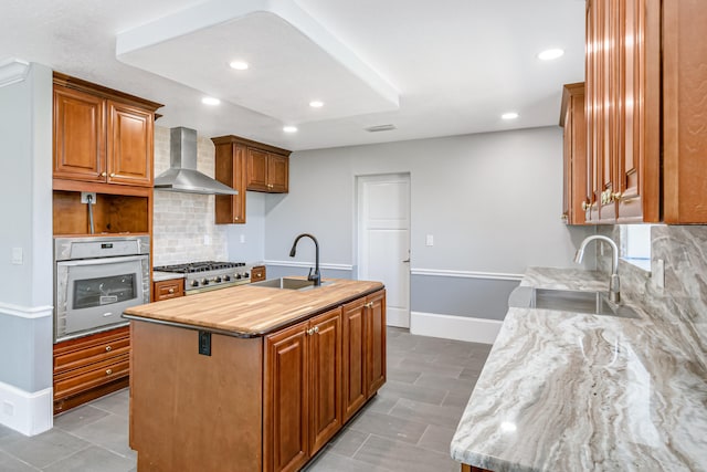 kitchen featuring backsplash, white oven, wall chimney exhaust hood, and an island with sink