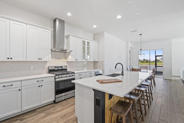 kitchen with white cabinetry, sink, wall chimney range hood, stainless steel gas range, and a center island with sink