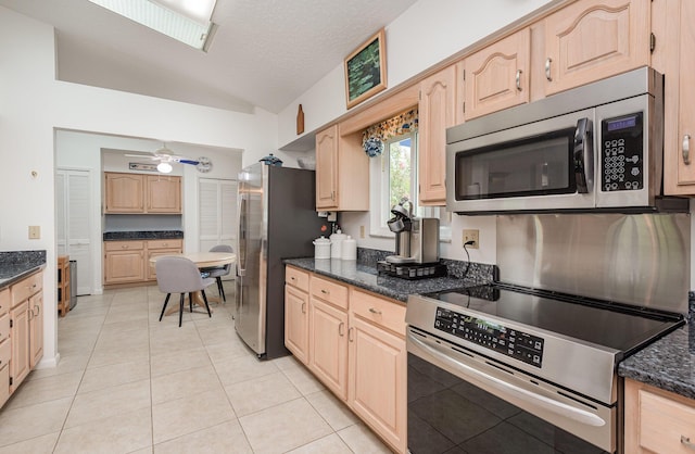 kitchen with light tile patterned flooring, lofted ceiling, light brown cabinetry, dark stone countertops, and appliances with stainless steel finishes