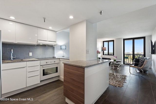 kitchen with kitchen peninsula, decorative backsplash, white oven, stone countertops, and white cabinets