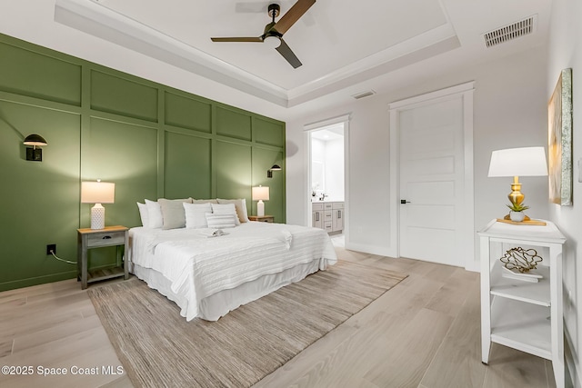 bedroom with ceiling fan, light hardwood / wood-style floors, a raised ceiling, and ensuite bath