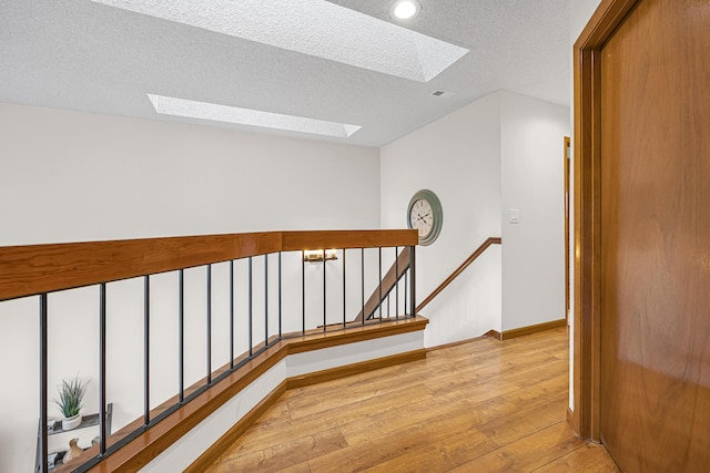 corridor with a skylight, light wood-style flooring, a textured ceiling, and an upstairs landing