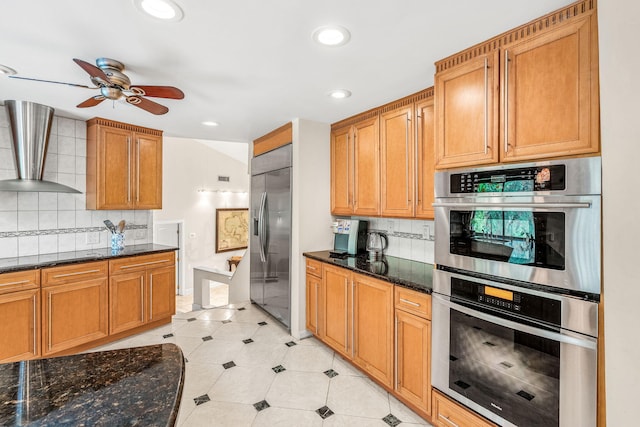 kitchen featuring wall chimney range hood, ceiling fan, appliances with stainless steel finishes, backsplash, and dark stone counters